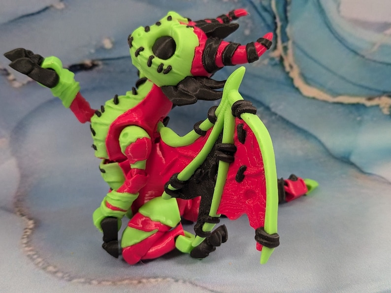 May include: A 3D-printed dragon figurine in vibrant colors. The dragon features a lime green head, limbs, and tail, contrasted by a red body and wings. Black accents highlight the horns, claws, and wing details. The dragon is posed on a blue and white surface.