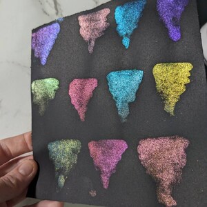 Pu&ograve; includere: Una superficie nera con 12 campioni di vernice iridescente in vari colori, tra cui turchese, viola, rosa e oro. I campioni sono disposti in una griglia.
