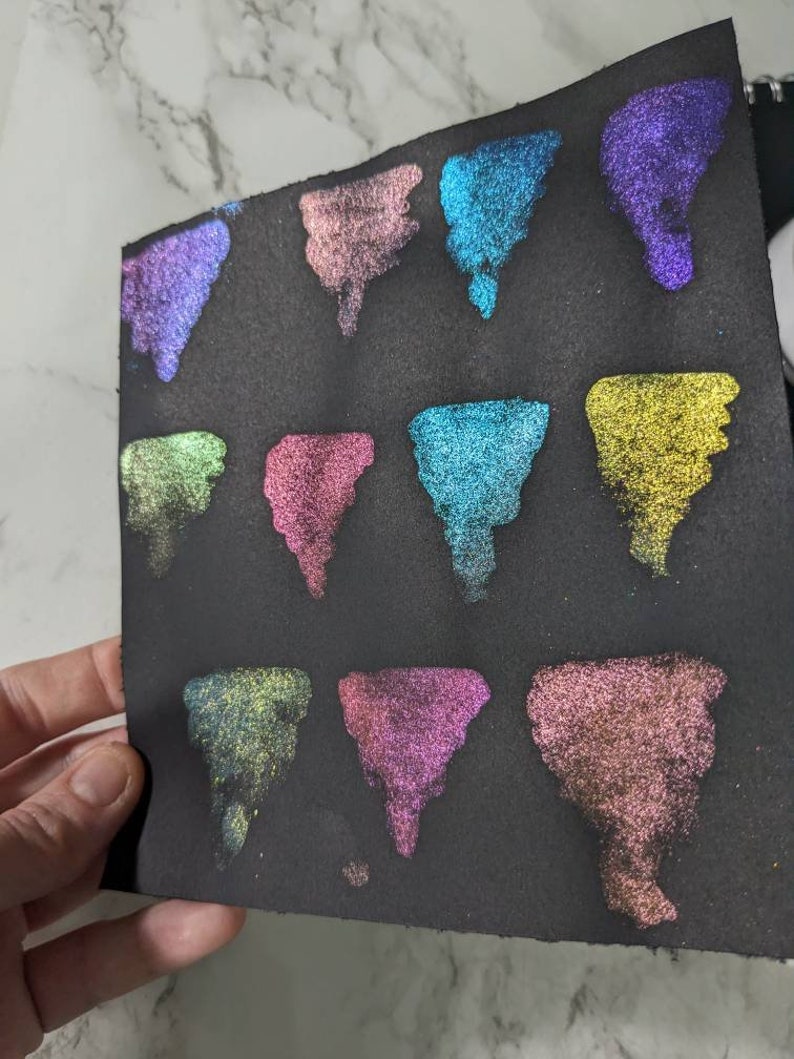 Pu&ograve; includere: Una superficie nera con 12 campioni di vernice iridescente in vari colori, tra cui turchese, viola, rosa e oro. I campioni sono disposti in una griglia.