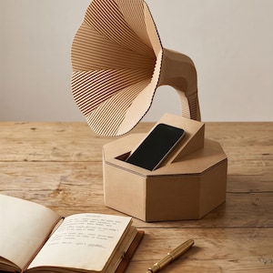 Può includere: Un altoparlante per telefono a forma di grammofono in cartone. L'altoparlante ha un grande corno scanalato e una base esagonale. Uno smartphone è inserito in una fessura sulla base. Un taccuino aperto e una penna sono sulla superficie di legno.