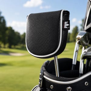 May include: A black textured golf putter headcover with white piping trim and a small black rectangular tag featuring a white crown logo. The headcover is attached to a metal putter shaft resting inside a black golf bag with silver metal grommets.