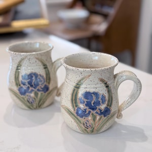 May include: Two ceramic mugs with a speckled cream glaze, each featuring a hand-painted blue iris flower with green leaves. The mugs have a rounded body, a wide rim, and a curved handle on the side, showcasing a consistent floral design.