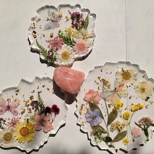 May include: Three clear resin coasters with a scalloped edge, each featuring pressed flowers and butterflies. The coasters are decorated with gold leaf and a pink rose quartz crystal is in the centre.