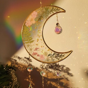 May include: A crescent moon-shaped suncatcher featuring a gold metal frame filled with clear resin, embedded with pressed pink flowers, green ferns, and gold leaf flakes. It hangs from a gold chain with multiple iridescent crystal prisms.