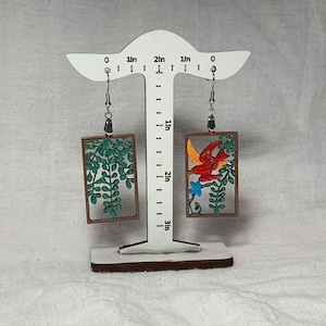 May include: A pair of wooden earrings with a brown frame. One earring features a green leafy design, while the other features a red bird with yellow accents and blue flowers.