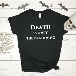 May include: Black t-shirt with the text "DEATH IS ONLY THE BEGINNING" in white. The shirt is styled on a white wooden surface with Halloween decorations, including a skull, skeleton hand, and bat garland.