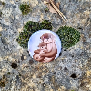 May include: A blue and white button with a cartoon image of two otters hugging. The larger otter is brown and has a pink nose. The smaller otter is brown and has a pink nose. The otters are on a light blue background.