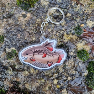 Pode incluir: Um chaveiro de acrílico transparente com uma ilustração de desenho animado de um peixe vermelho e marrom com contorno branco. O peixe tem uma boca grande e barbatanas vermelhas. O chaveiro está preso a um anel de chave de metal prateado.