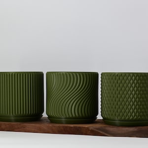 May include: Three green ceramic planters with different embossed patterns. The planters are arranged on a wooden surface.