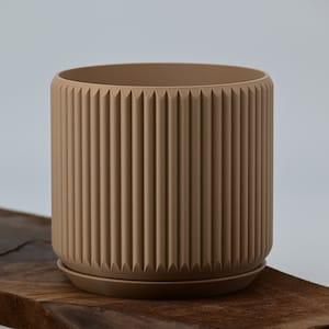May include: A light brown ceramic planter with a ribbed design and a matching saucer. The planter is sitting on a wooden surface.