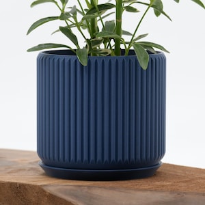 May include: A navy blue, cylindrical planter with vertical ribbed detailing. The planter includes a matching saucer and holds a green leafy plant. The pot is set on a wooden surface against a white background. A stylish addition to any home decor.