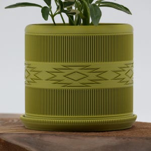 May include: A chartreuse-colored cylindrical planter with a geometric design and vertical ribbed texture. The planter includes a matching saucer and holds a small green plant. The design features a band of diamond shapes. The planter is approximately 6 inches tall.