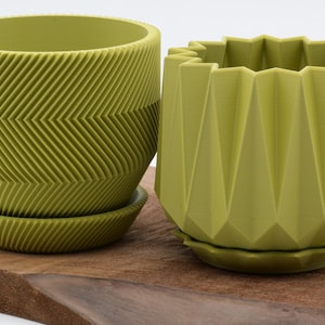 May include: Two olive green ceramic planters with matching saucers. One planter has a chevron pattern, the other has a geometric, faceted design. Both are on a wooden surface.