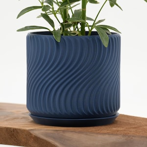 May include: A navy blue ceramic planter with a wavy, textured design. The cylindrical pot sits on a matching saucer and holds a green plant. The planter is displayed on a wooden surface against a white background.