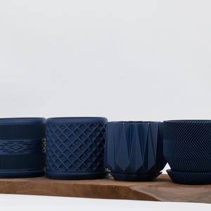 May include: Four navy blue planters with textured designs sit on a wooden board. The planters have various geometric patterns, including a diamond grid, chevron, and a tribal-inspired design. Each planter has a matching saucer.