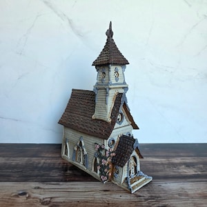 May include: A ceramic church figurine featuring a tall steeple, a brown shingled roof, and off-white siding. The structure includes arched windows with blue trim, a small entrance, and decorative pink floral vines climbing up the side wall.
