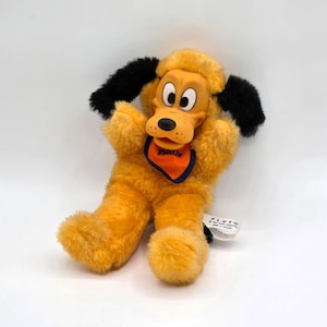 May include: A plush Pluto toy featuring golden yellow fur, long black ears, and a molded plastic face. The character wears an orange neckerchief with a dark blue border and the name Pluto printed on a white fabric tag attached to the side.