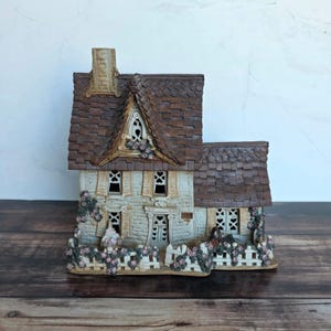 May include: A ceramic house figurine featuring a textured brown shingled roof, a cream-colored brick facade, and a white picket fence adorned with sculpted pink flowers. The structure includes multiple windows with heart-shaped cutouts and a chimney.