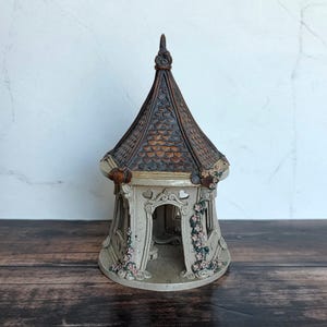 May include: A ceramic gazebo figurine featuring a dark brown, shingled, conical roof and a light beige, open-walled structure. The exterior is decorated with sculpted pink floral vines climbing the pillars and ornate, arched architectural details.