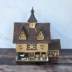 May include: A detailed ceramic house with a brown shingled roof, decorative windows, and a small steeple. The house has a light tan exterior with cut-out windows and a small barn area with miniature animal figurines. The house is approximately 8 inches tall.
