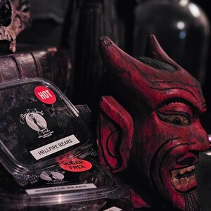 May include: Two clear plastic containers of black gummy candies with labels that say "Hellfire Bears" and "Monster Bears". The container labeled "Hellfire Bears" has a red sticker that says "HOT". The container labeled "Monster Bears" has a red sticker that says "Sugar Free". A red wooden devil mask is in the background.