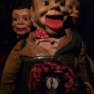 May include: A vintage ventriloquist dummy with a red tongue and a pink polka dot bow tie. The dummy is holding a clear container of "Psycho Pineapple" treats. Two other dummies are in the background. The treats are from "Davis' Spooky Treats".