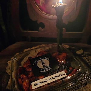 May include: A clear plastic container filled with mango-flavored candy. The container has a black and white label that says "Treats" with a witch illustration and the text "daily spooky treats @instagram". The label also says "Mango Madness".