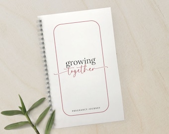 May include: A white spiral bound journal with a red border around the cover. The cover has the text "growing together" in black cursive writing and the text "Pregnancy Journey" in black print.