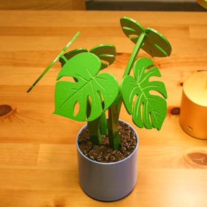 May include: A decorative artificial monstera plant in a light blue pot, with bright green leaves. A gold-colored desk lamp with a round top is next to the plant. The items are on a wooden surface.