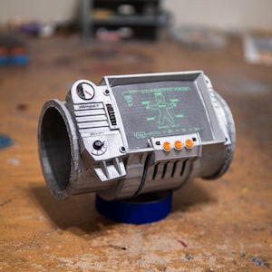 May include: A silver-colored cylindrical wrist-mounted device featuring a rectangular screen with a green graphic of a cartoon figure. The unit includes a small circular gauge, a black dial, and three orange circular buttons on the front panel.