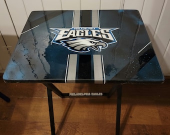 Puede incluir: Una mesa plegable de metal negro con una tapa de mesa azul, negra y plateada con el logotipo de los Philadelphia Eagles y las palabras "Philadelphia Eagles".