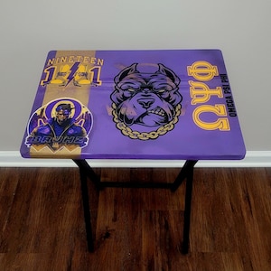 May include: A purple folding table with black legs. The tabletop features a gold stripe with the text "NINETEEN 1941" and a graphic of a man. A bulldog graphic with a gold chain and the Greek letters "ΦΨΩ" are also on the table.