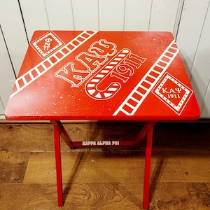 May include: Red folding table with white painted design. The design includes the letters "KAY" and "1911" and the Greek letters "ΚΑΨ". The table has the words "Kappa Alpha Psi" printed on the bottom.