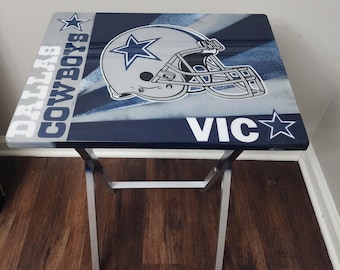 Op de afbeelding: Vierkante klaptafel met een Dallas Cowboys-thema. Het tafelblad toont een afbeelding van een voetbalhelm en de woorden "DALLAS COWBOYS" en "VIC" in het wit. De tafel heeft een zilverkleurig metalen frame.