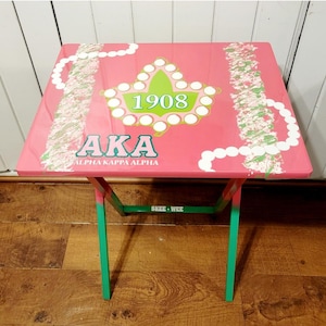 May include: A pink folding table with a green frame. The table top features a green and white sorority symbol with the year 1908 and the letters AKA, which stands for Alpha Kappa Alpha.