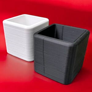May include: Two square planters, one white and one black, are set against a red backdrop. The planters have a textured, layered appearance. The white planter is on the left, and the black planter is on the right.