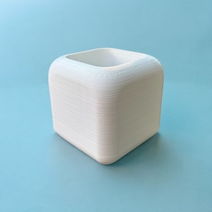 Könnte beinhalten: Ein weißer, quadratischer Pflanzer mit abgerundeten Kanten. Der Pflanzer besteht aus einem glatten, strukturierten Material und hat einen leicht erhöhten Rand oben.