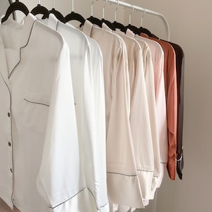 May include: A rack of eight long-sleeved shirts in various shades of white, pink, and brown. The shirts have black trim around the collar and cuffs.