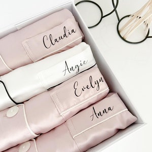 May include: A white box containing a pink satin pyjama set. The pyjamas are folded and stacked on top of each other. Each pyjama set has a different name embroidered on the collar: Claudia, Angie, Evelyn, and Anna.