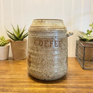 May include: A cylindrical ceramic canister with a speckled beige and brown finish. The word COFFEE is embossed on the front. It features a slightly tapered top, a small side handle, and horizontal ridges along the lower body of the vessel.