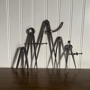 May include: A collection of antique metal calipers of varying sizes, arranged on a wooden surface against a white paneled wall. The calipers are a dark, aged metal, showing signs of wear and use. The tools have pointed tips and adjustable arms.