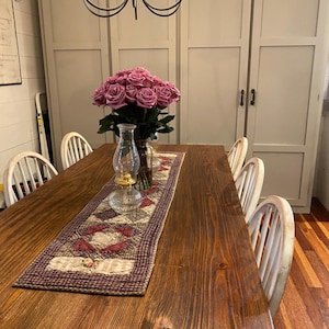 May include: A rustic wooden dining table with a floral centerpiece of pink roses in a glass vase. A decorative quilted table runner with red and cream patterns runs down the center. A vintage glass oil lamp sits on the runner, and white chairs surround the table.