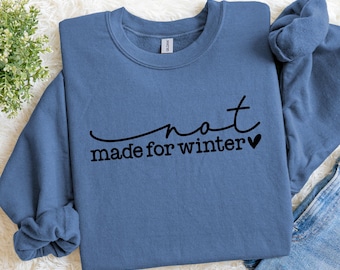 Puede incluir: Sudadera azul con cuello redondo y la frase "not made for winter" en negro. La sudadera está doblada, con una parte de unos vaqueros a la vista.