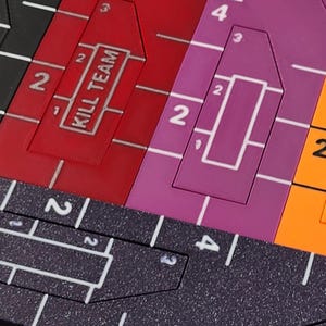 Op de afbeelding: Close-up van een kleurrijk spelbord met geometrische vormen en cijfers. Het bord heeft rode, paarse en oranje secties met witte rasterlijnen en de tekst "KILL TEAM".