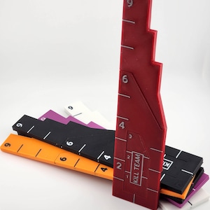 May include: A collection of colorful, geometric game measuring tools. The set includes rulers in red, orange, black, purple, and white. Each ruler is marked with white numerical increments. The red ruler has the text "KILL TEAM".