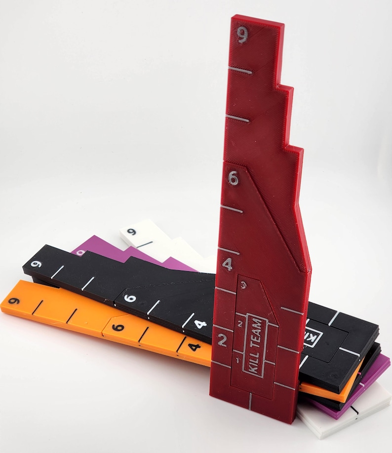 Op de afbeelding: Een verzameling kleurrijke, geometrische spelmeetinstrumenten. De set bevat linialen in rood, oranje, zwart, paars en wit. Elke liniaal is gemarkeerd met witte numerieke stappen. De rode liniaal heeft de tekst "KILL TEAM".