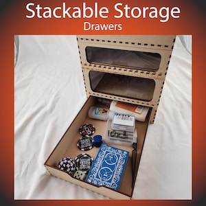 May include: A wooden stackable storage drawer unit with three drawers. The top two drawers are clear and the bottom drawer is open, revealing various items including dice, game pieces, and small containers.