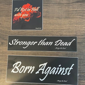 May include: Three black and white stickers with gothic-style lettering. The first sticker reads "I'd Rot in Hell with you" with a red rose in the background. The second sticker reads "Stronger than Dead". The third sticker reads "Born Against". All three stickers have the text "Amigo the Devil" at the bottom.