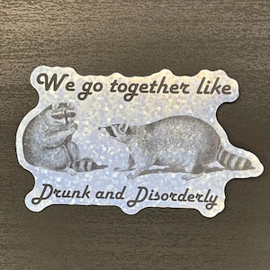 May include: A sticker with a glittery blue background featuring two raccoons and the text "We go together like drunk and disorderly".