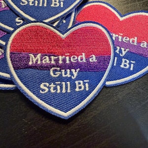 May include: Heart-shaped embroidered patches in the colours of the bisexual pride flag: pink, purple, and blue. The text "Married a Guy Still Bi" is embroidered in white on the blue section of the patch. Several patches are visible.